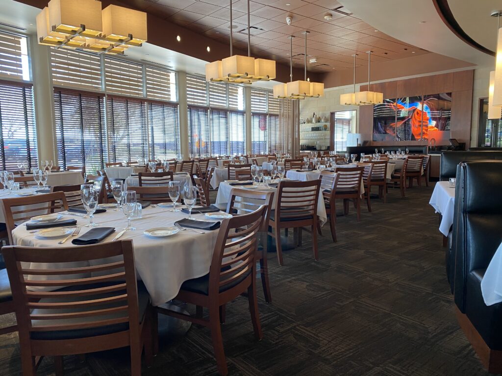 A spacious dining area at Ocean Prime Restaurant Phoenix features neatly arranged wooden tables and chairs. Tables are set with white tablecloths, glasses, plates, and cutlery. Large windows with blinds allow natural light, and modern pendant lights hang from the ceiling.