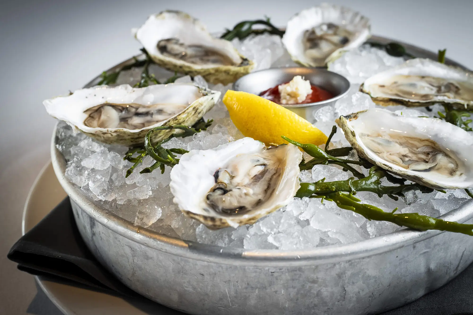 A metal tray filled with crushed ice holds six opened oysters with seaweed garnishes, centered around a small dish of cocktail sauce with horseradish and a lemon wedge. The tray is set on a white plate, and a dark cloth napkin is folded underneath.