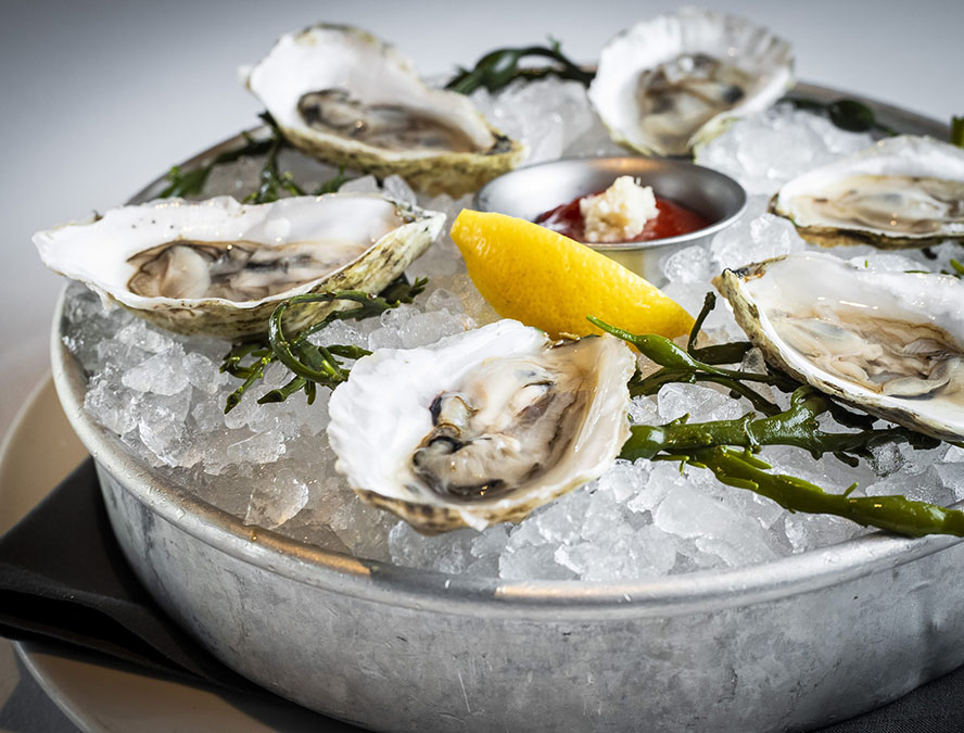 A metal tray filled with crushed ice holds six opened oysters with seaweed garnishes, centered around a small dish of cocktail sauce with horseradish and a lemon wedge. The tray is set on a white plate, and a dark cloth napkin is folded underneath.