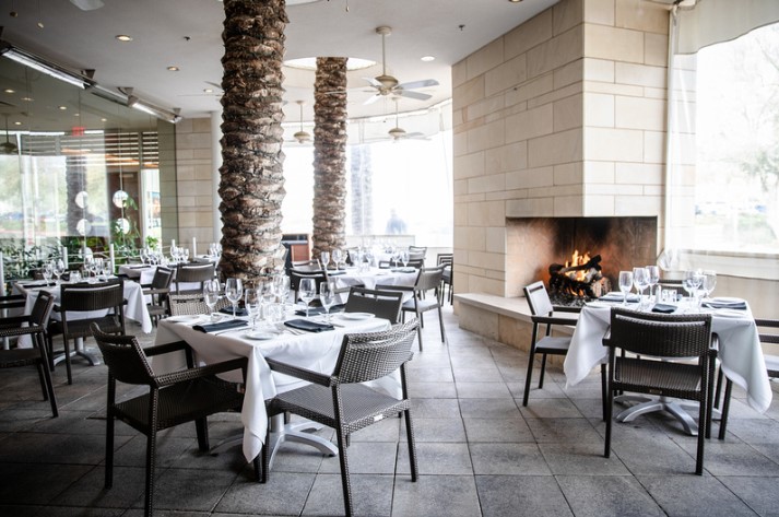 Ocean Prime Restaurant Phoenix interior with tables set for dining, complete with white tablecloths, glassware, and cutlery. The space features palm tree columns, large windows allowing natural light, ceiling fans, and a cozy fireplace on the right.